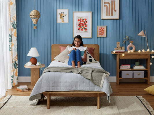 A cosy bedroom with a wooden bed, light grey quilt, and green throw. Blue panelled wall features framed art. A child sits on the bed.