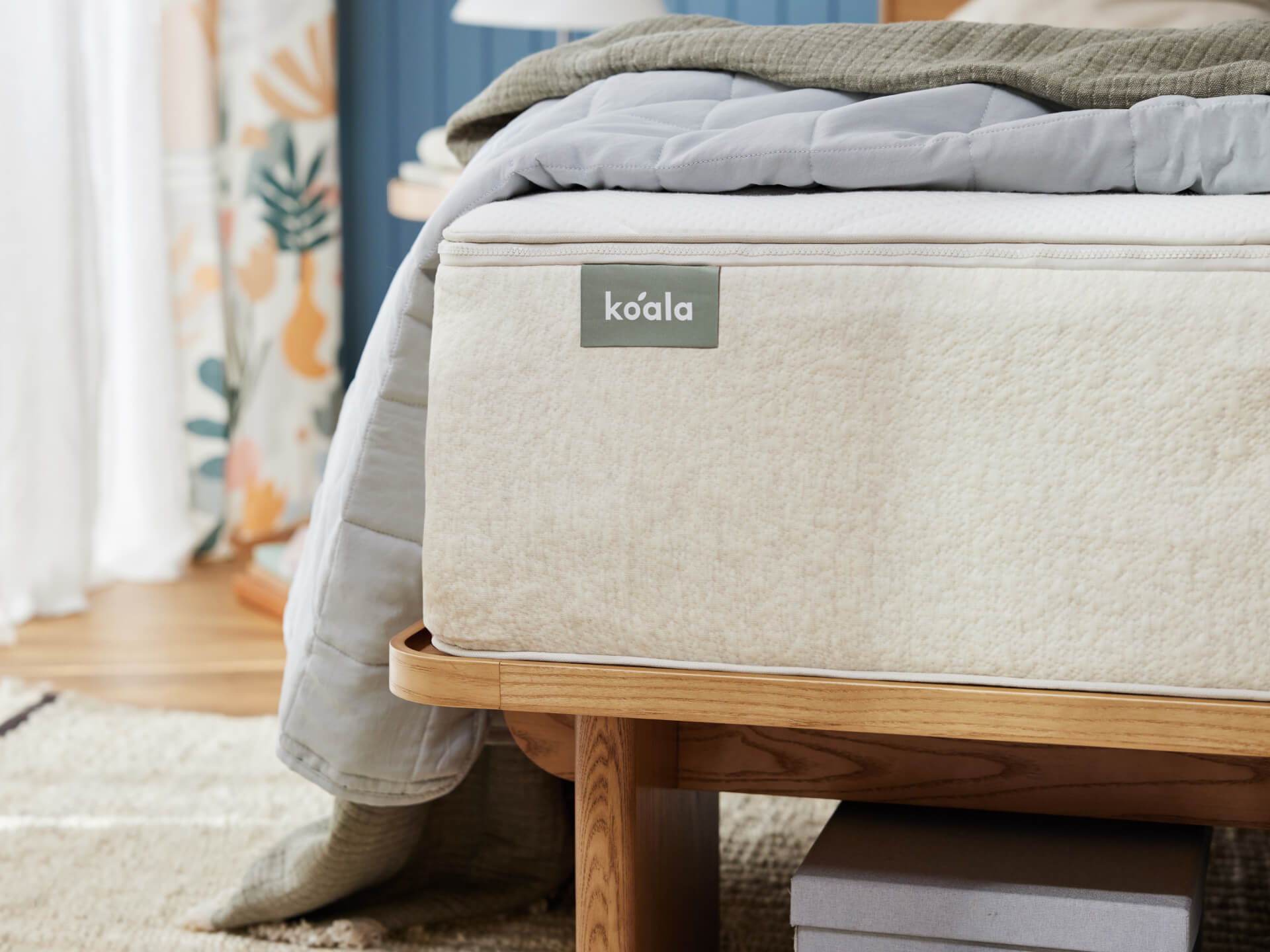 Beige mattress on a wooden bed frame, with a grey blanket on top. Floral curtains and a light-coloured rug in the background.
