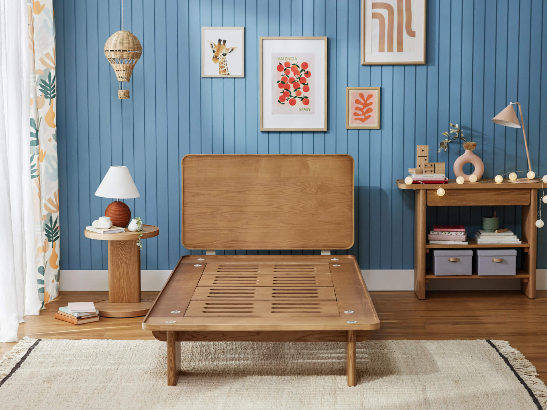 Wooden bed frame with slatted base, light wood side table and desk, blue panelled wall, and framed art. Cream curtain and rug.