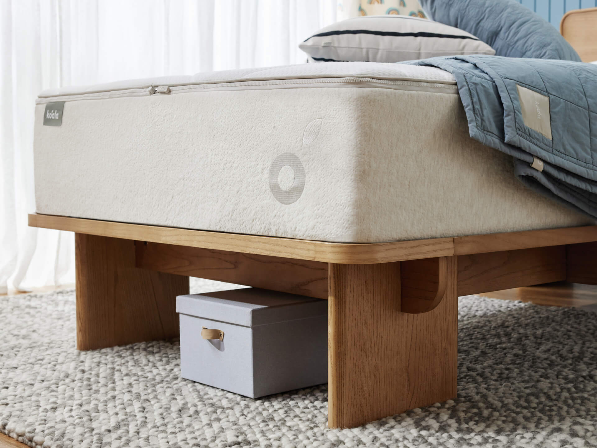 Wooden bed frame with light grey mattress on a textured grey rug. A blue quilt partially covers the bed, with a small box underneath.