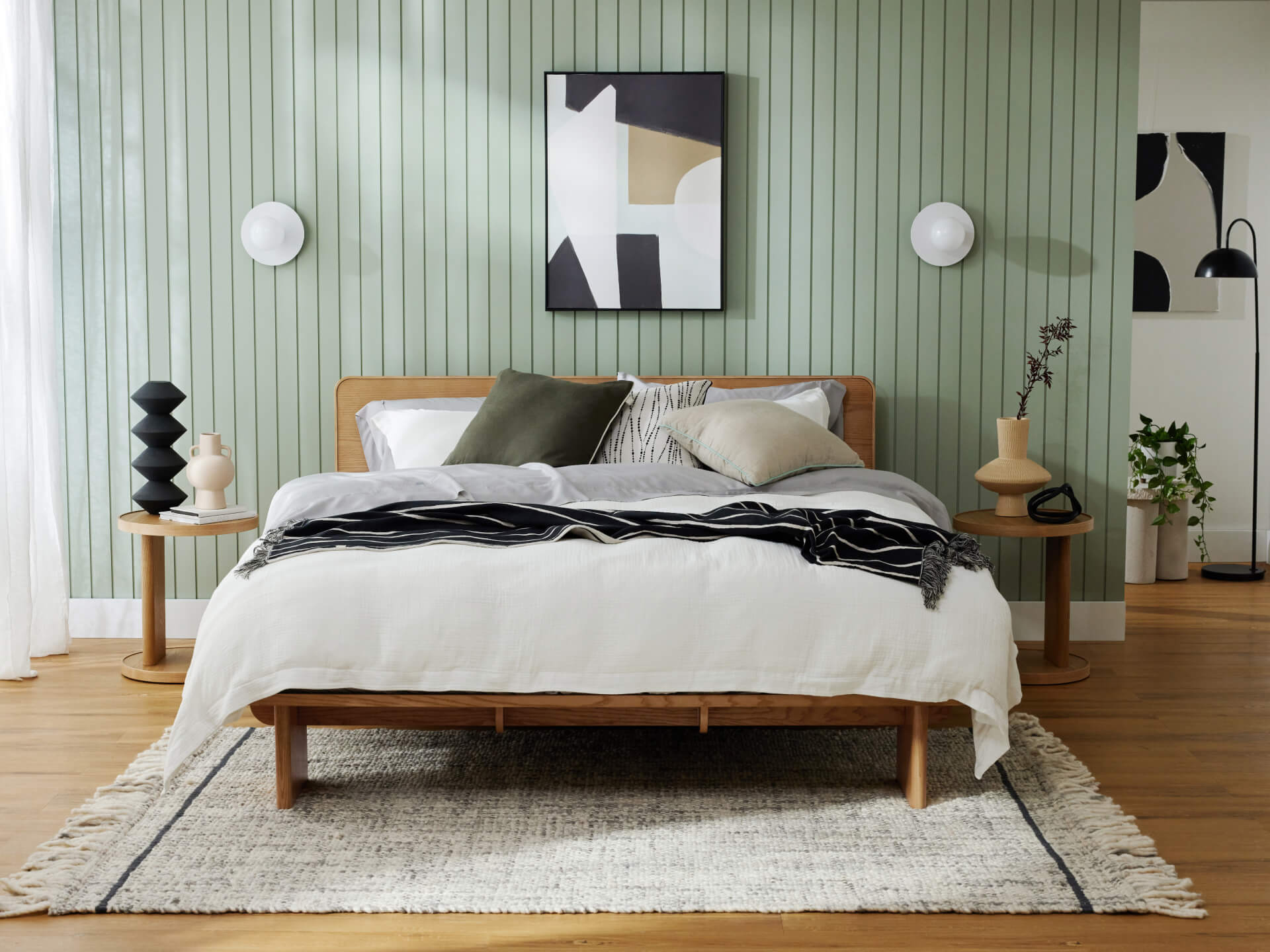 Minimalist bedroom with a wooden bed, white bedding, and green and grey cushions. Light green panelled wall, abstract art, and round bedside tables.