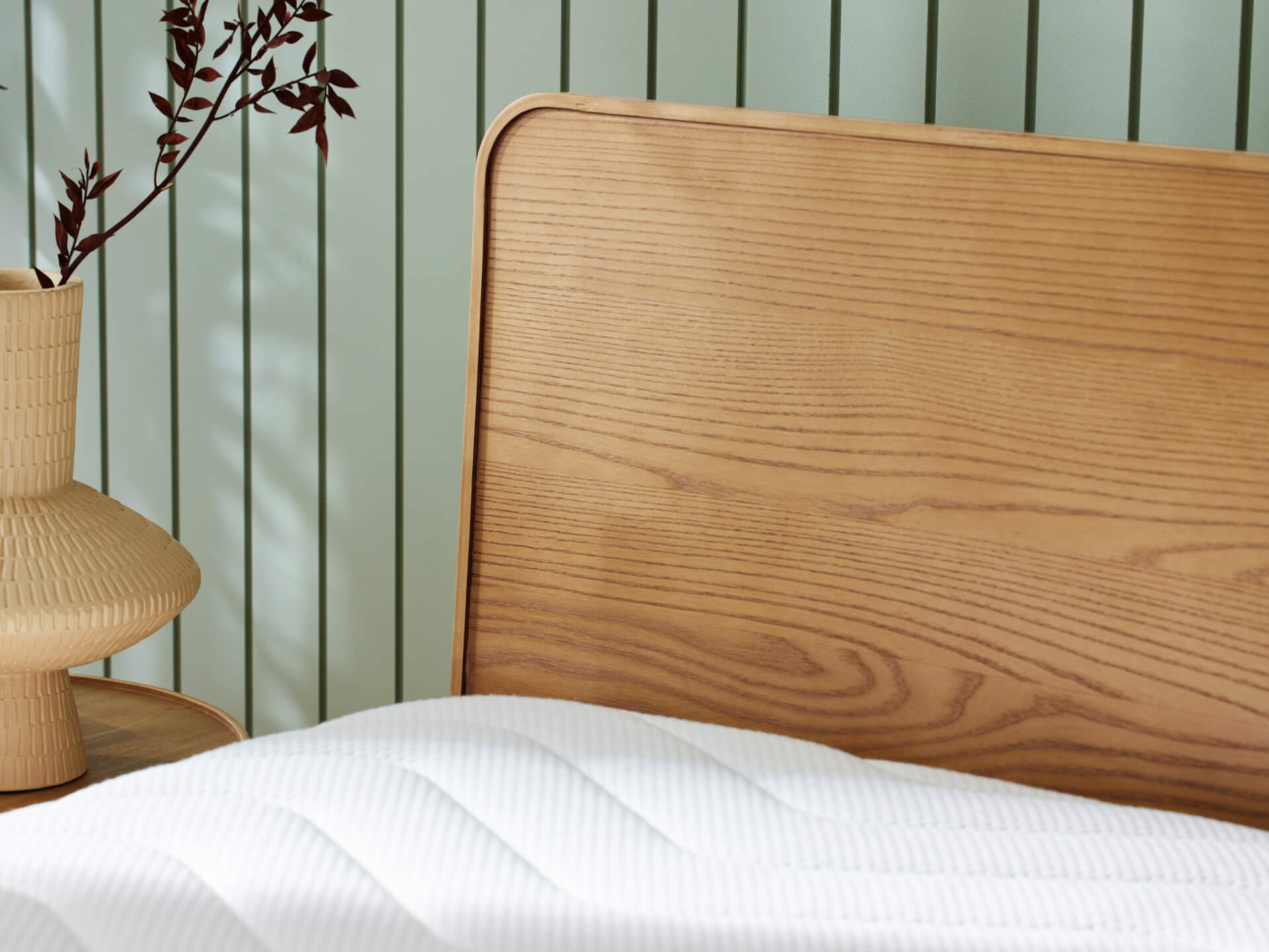 Wooden bedhead with light wood grain pattern, white quilted mattress, cream textured vase with dark red branches, against vertical pale green wall.