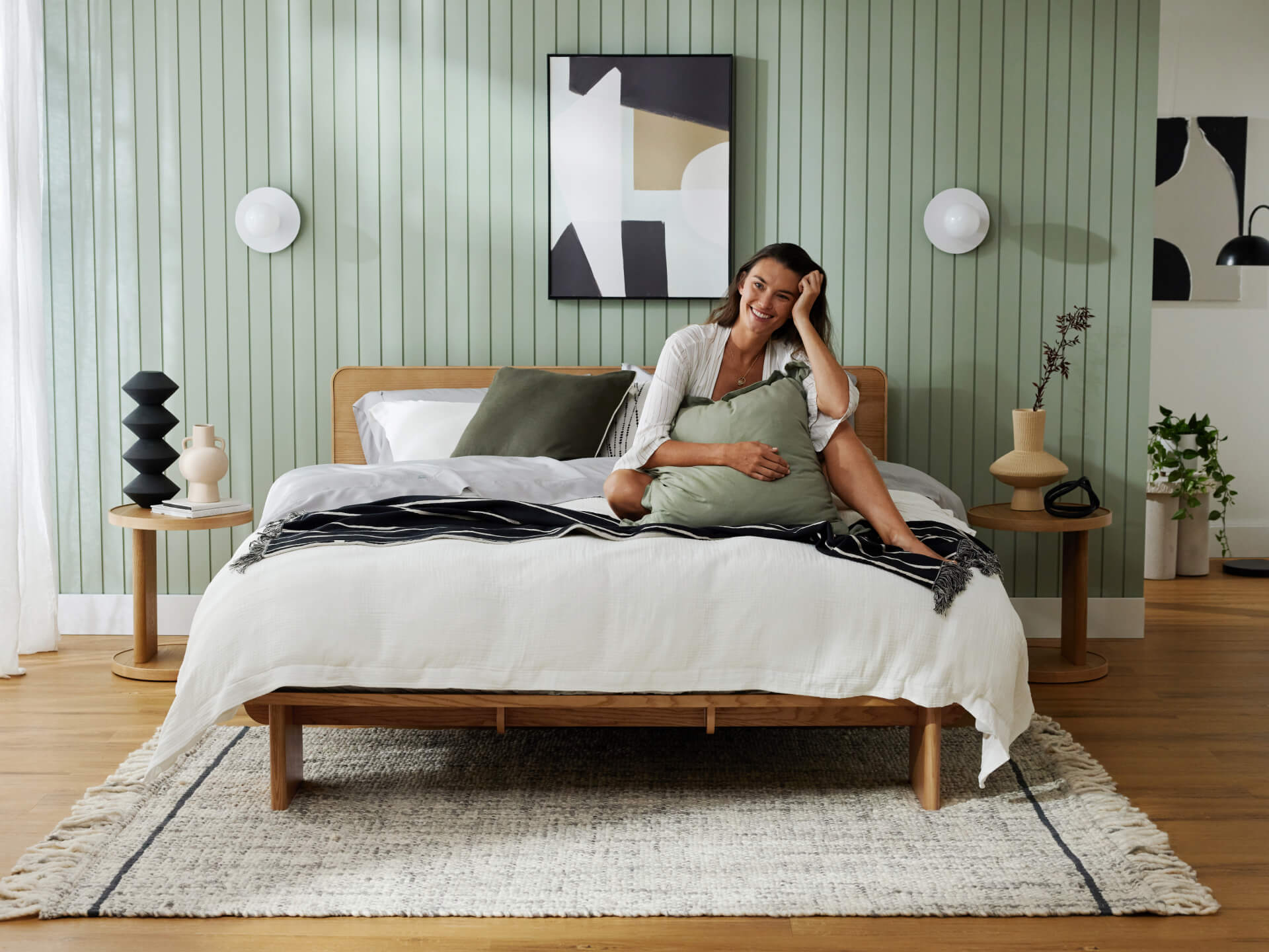 Bedroom with a wooden bedframe, white and grey bedding, light green cushions, and a grey rug on wooden flooring. Pale green panelled walls and abstract artwork.