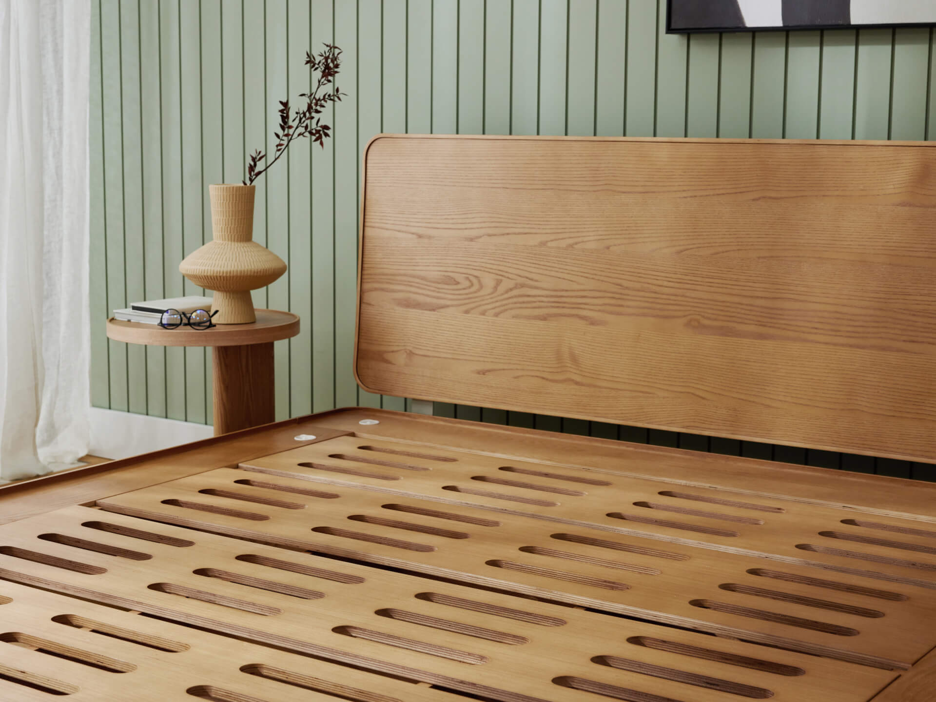 Wooden bed frame with slatted base, curved headboard. Nearby is a round side table with a vase, books, against light green panelled wall.