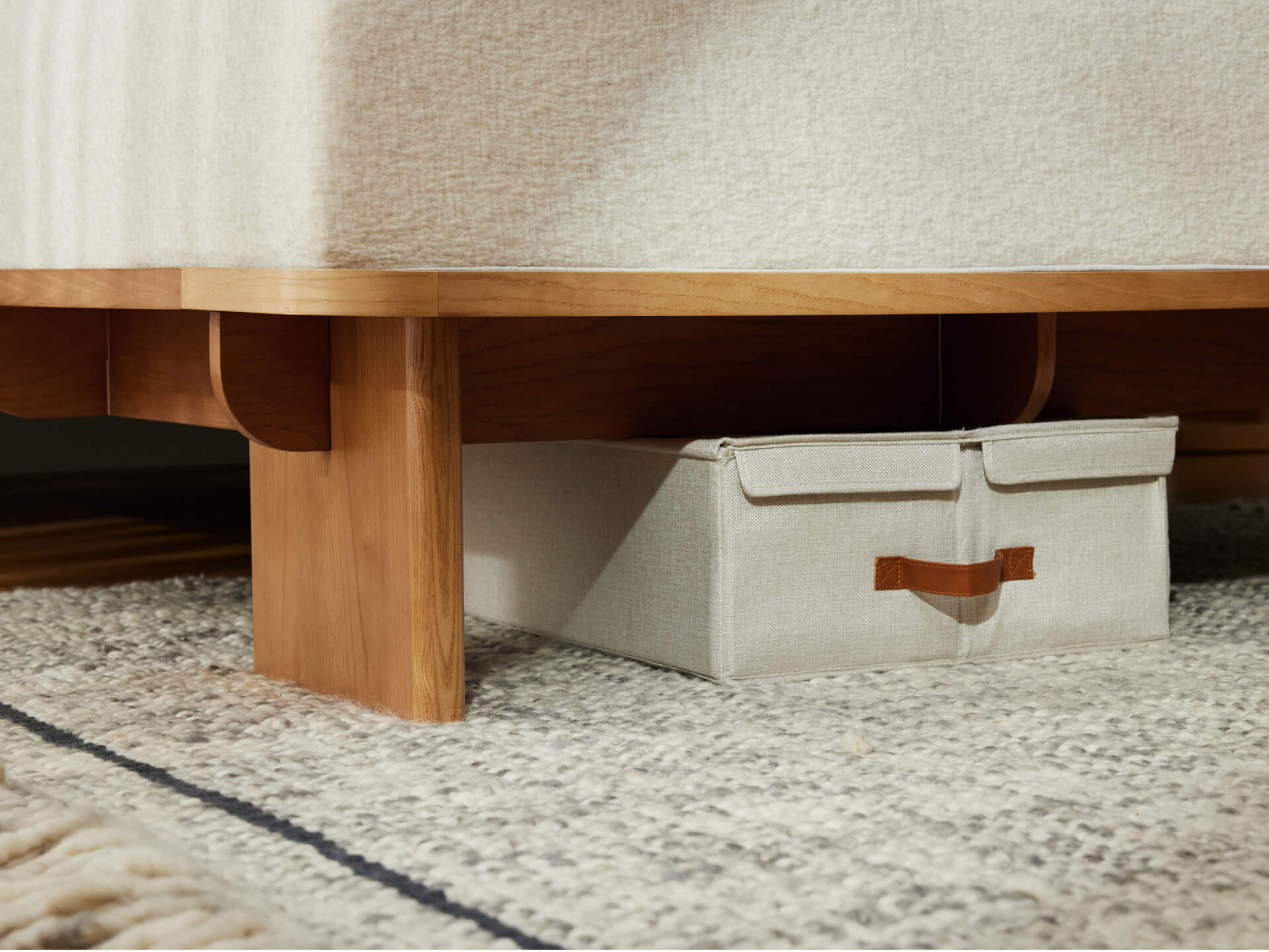 Light wood bed frame with beige upholstery, storage box underneath, and a textured rug in the foreground.