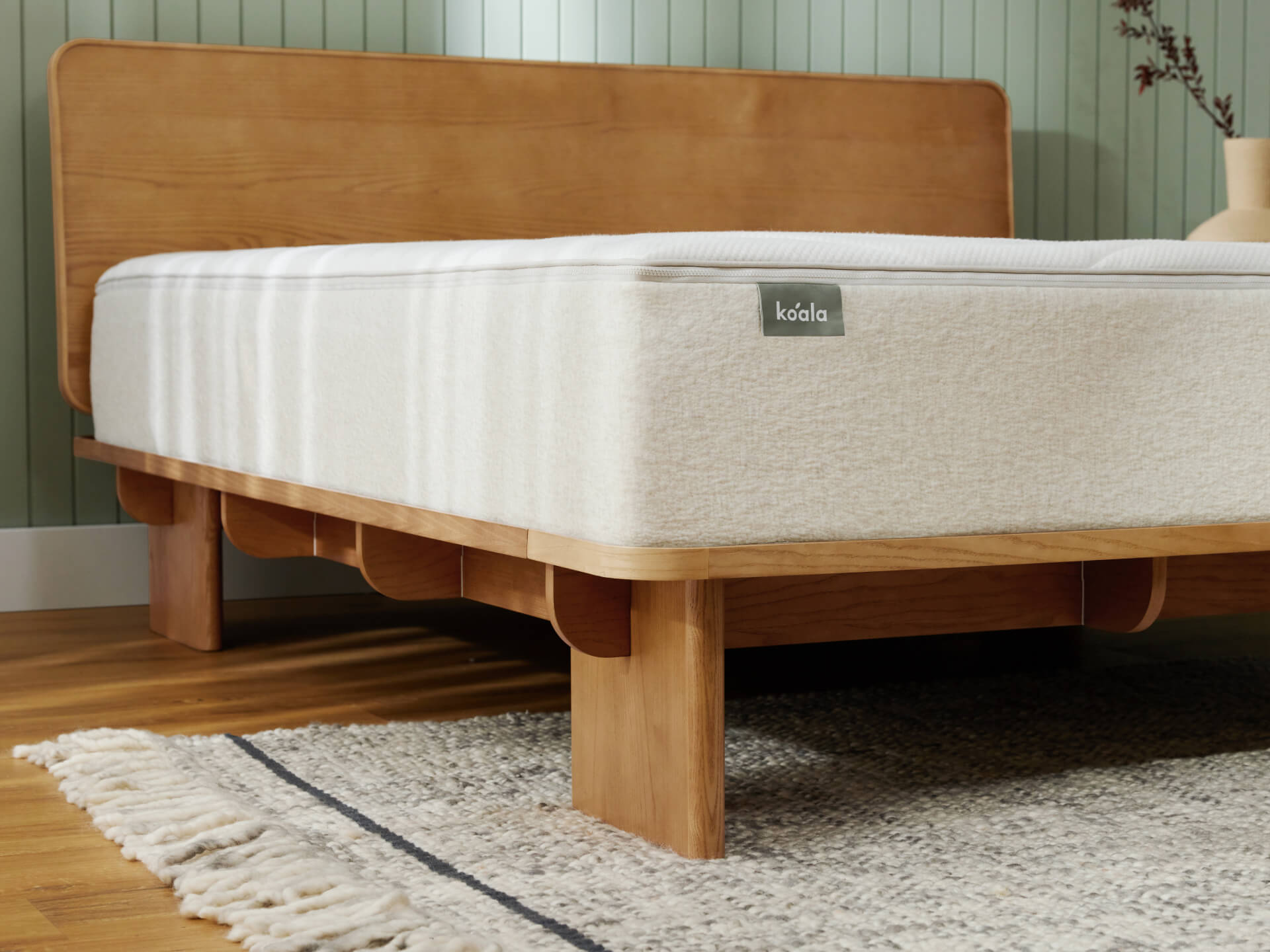 Wooden bed frame with light beige mattress on a grey rug. The room has green wall panelling and a ceramic vase in the corner.