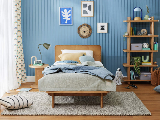 Cosy bedroom with light wood bed, blue sheets, and a tan headboard. Blue panelled wall, art, bookshelves, and rug complete the decor.