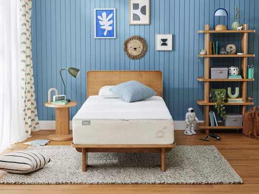 A cosy bedroom with a beige mattress on a wooden bed, blue throw pillow, light brown headboard, blue wall panel, shelf, lamp, and rug.