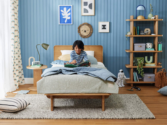 Child reading on a wooden bed with blue bedding in a cosy bedroom. Blue panelled wall with art, round side table, and tall shelf. Grey rug on floor.
