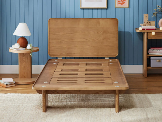 Wooden bed frame with headboard, set in a room with blue panelled walls. Nearby are a round side table with a lamp and a rectangular console.