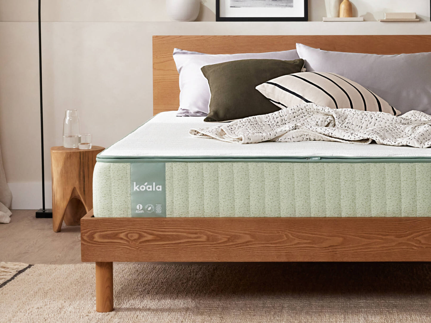 Wooden bed frame with a light green mattress and mixed pillows. Neutral-toned room with a rug, side table, and framed picture in the background.