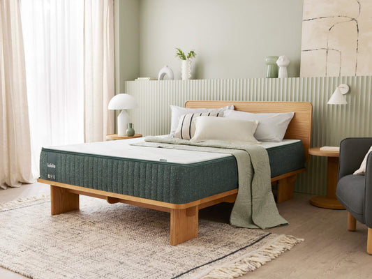 Modern bedroom with a green mattress on a wooden bed frame, light rug, and white bedding. Decor includes plants and lamps on a light green wall.