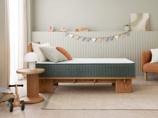 Children's bedroom with a single bed, green mattress, and wooden frame. A side table with a lamp, bunting on the wall, and a toy balance bike.