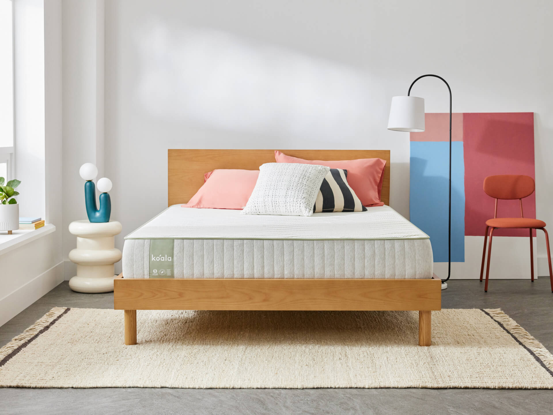 Modern bedroom with light wood bed frame, grey mattress, and coral pillows. Features a floor lamp, abstract wall art, and indoor plants.