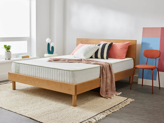 Wooden bed frame with light grey mattress, pink and white pillows, and a beige throw on a textured rug. Nearby are an orange chair and decor.