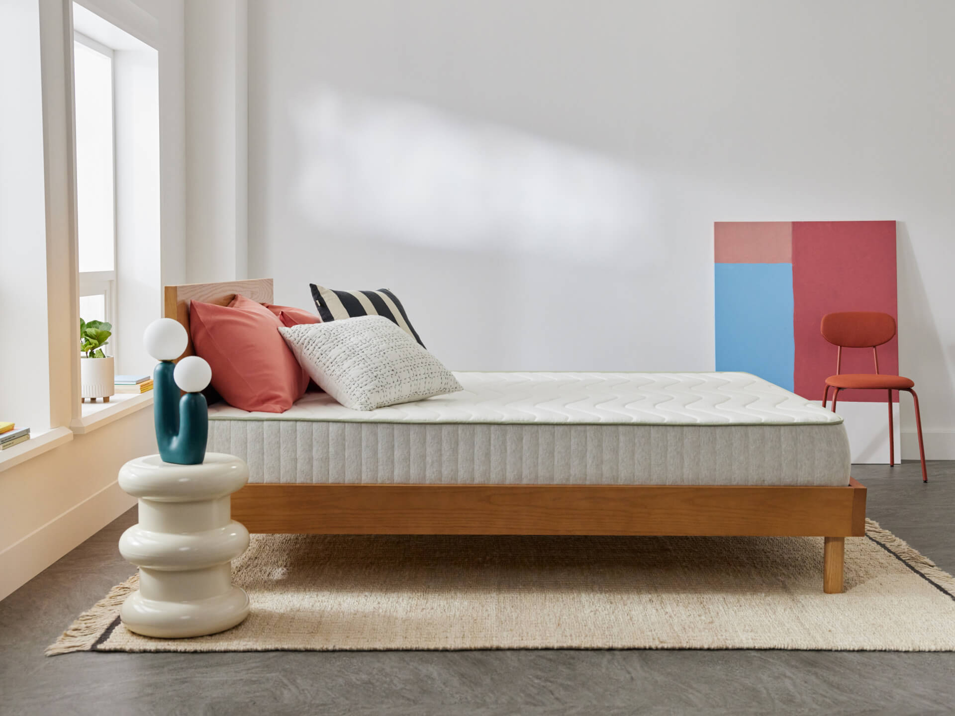 Minimalist bedroom with wooden bed frame and white mattress. Decorative cushions, white side table with lamp, and abstract art in the background.