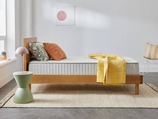 Minimalist bedroom with wooden bed frame, grey mattress, yellow quilt, decorative cushions, green side table, and pastel decor on a beige rug.
