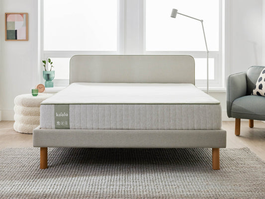 Modern bedroom with light grey bedframe and mattress, wooden legs, grey armchair, round ottoman, floor lamp, and light grey rug.