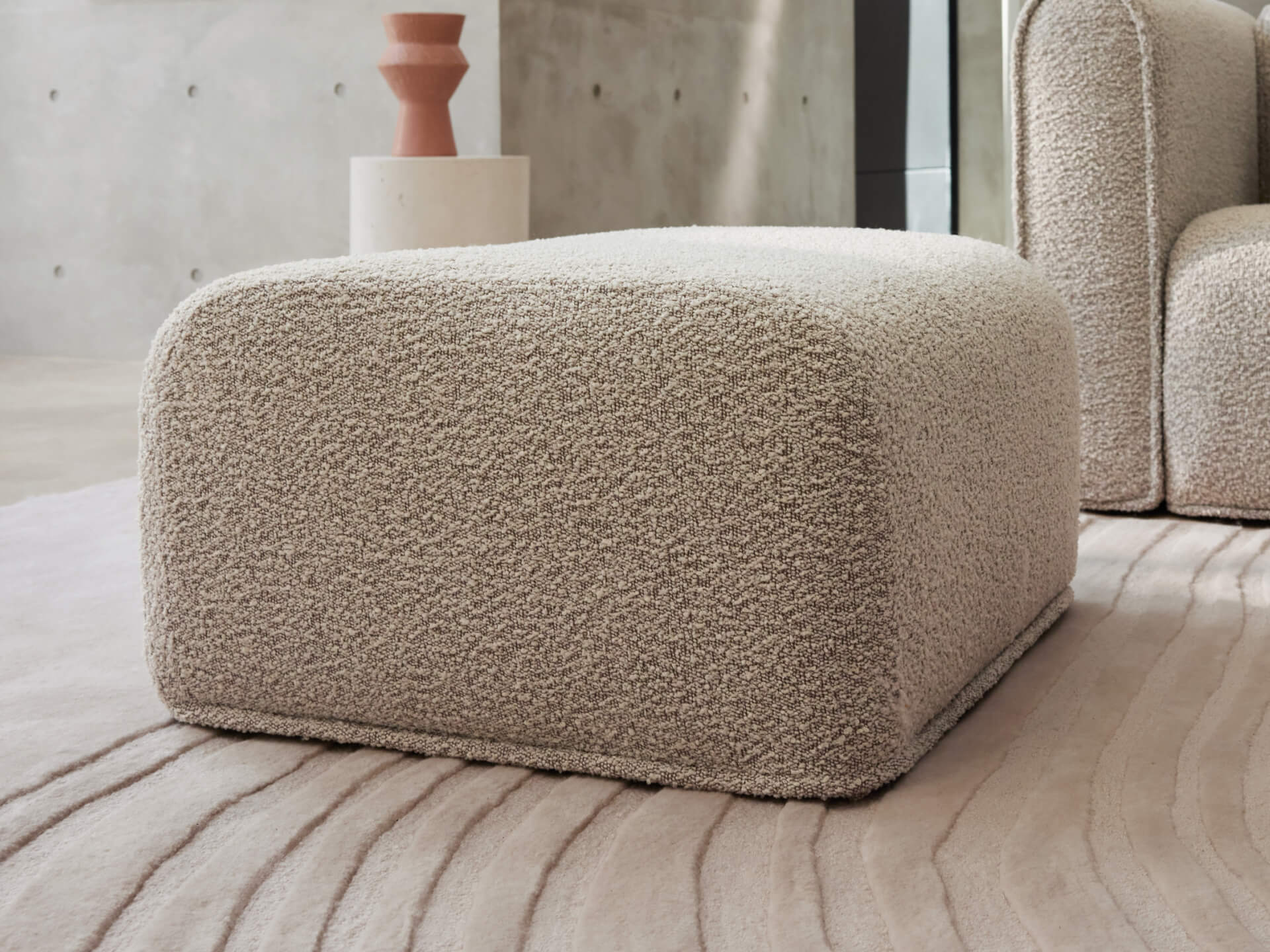 Beige boucle ottoman on a textured rug, next to a matching armchair. A brown vase on a white pedestal is in the background against a grey wall.