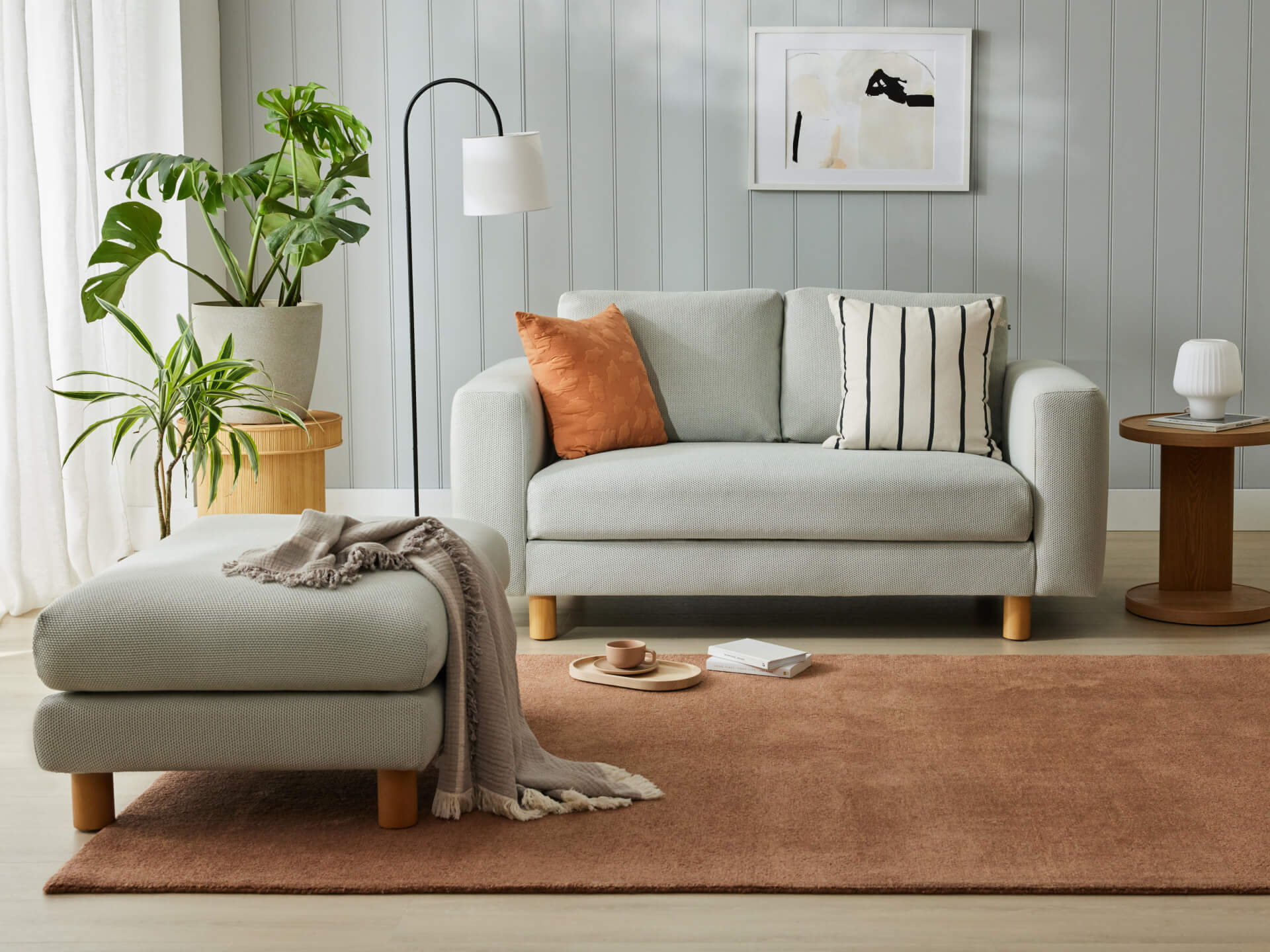 Light grey couch with an orange and striped cushion. Grey ottoman, brown rug, plants, and modern decor in a room with light green walls.