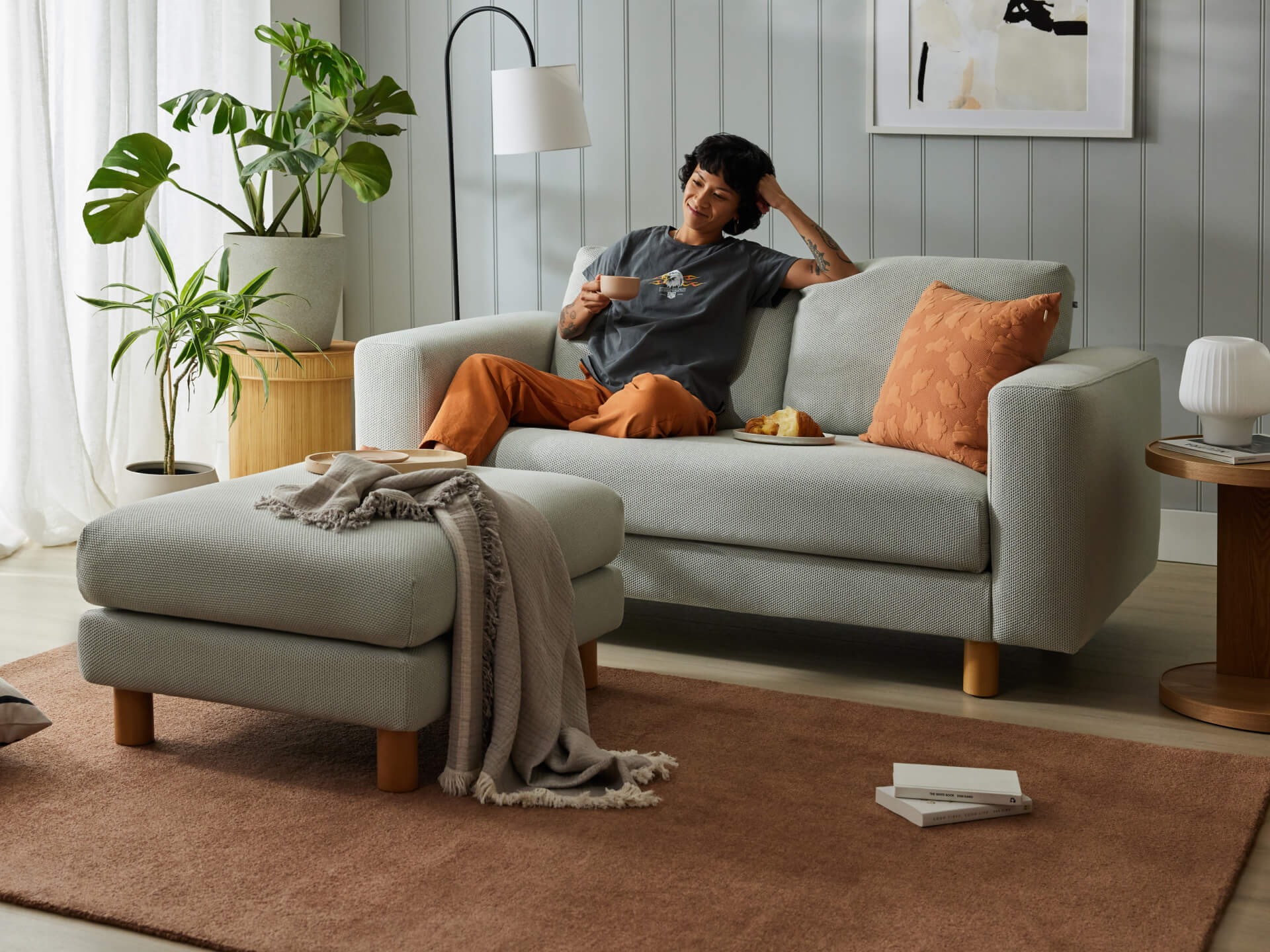 Cosy living room with light grey sofa and ottoman, terracotta cushion, green plants, and a floor lamp. A person is relaxing with a cup on the sofa.