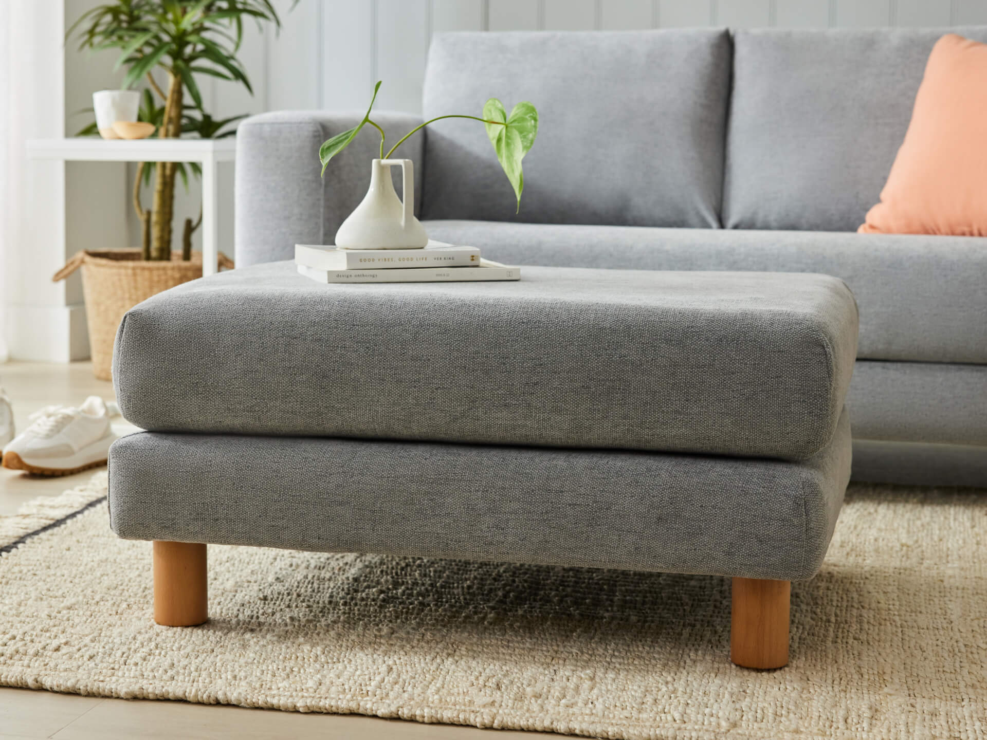 Grey upholstered ottoman with wooden legs on a cream rug, in front of a grey sofa. A white vase with a plant sits on top.