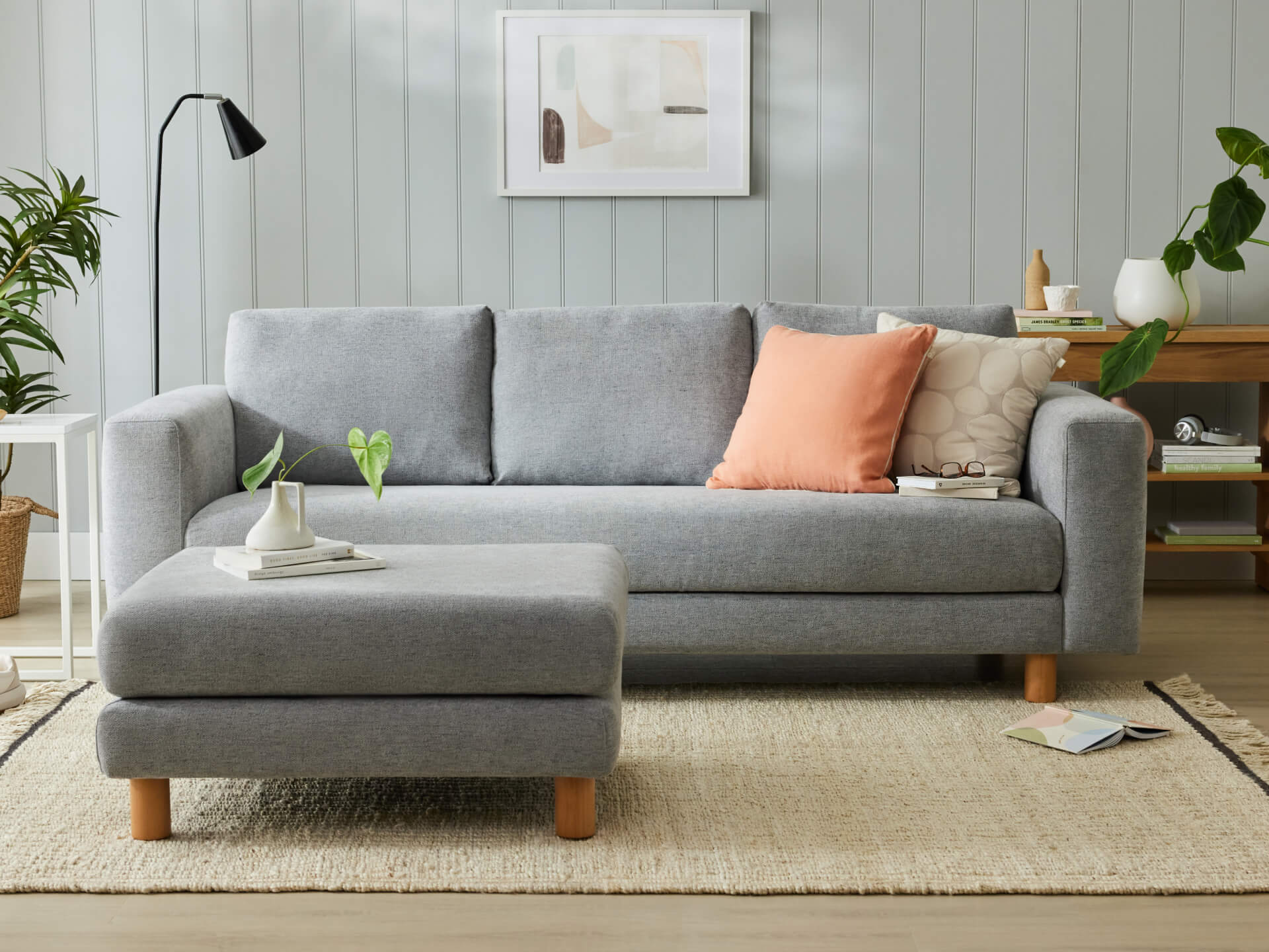 Light grey sofa with wooden legs, peach and beige cushions, in a living room with a matching ottoman, plants, a lamp, and wall art.