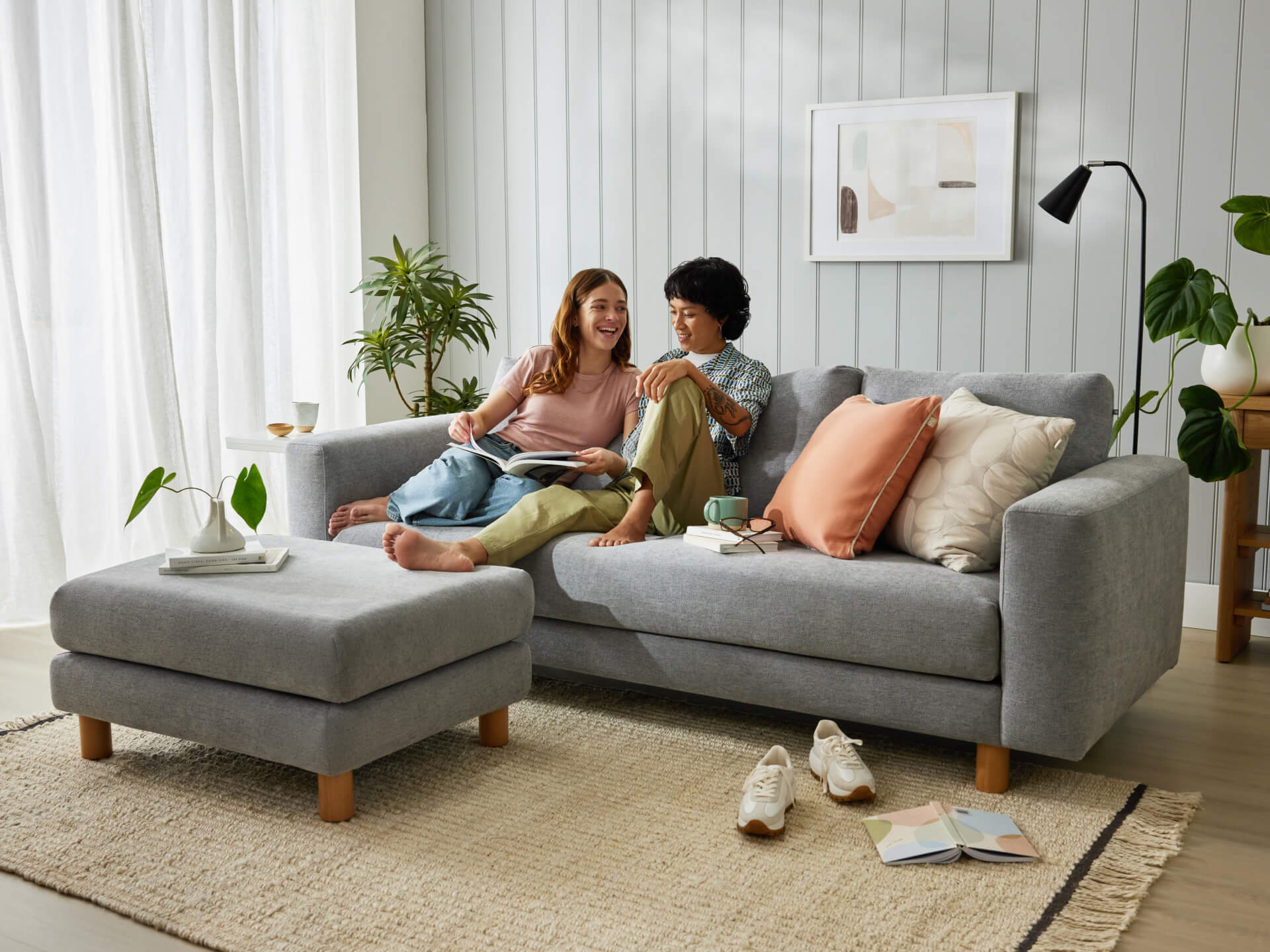 Couple relaxing on a light grey sofa with a matching ottoman, surrounded by plants in a cosy living room. Neutral decor accents and soft lighting.