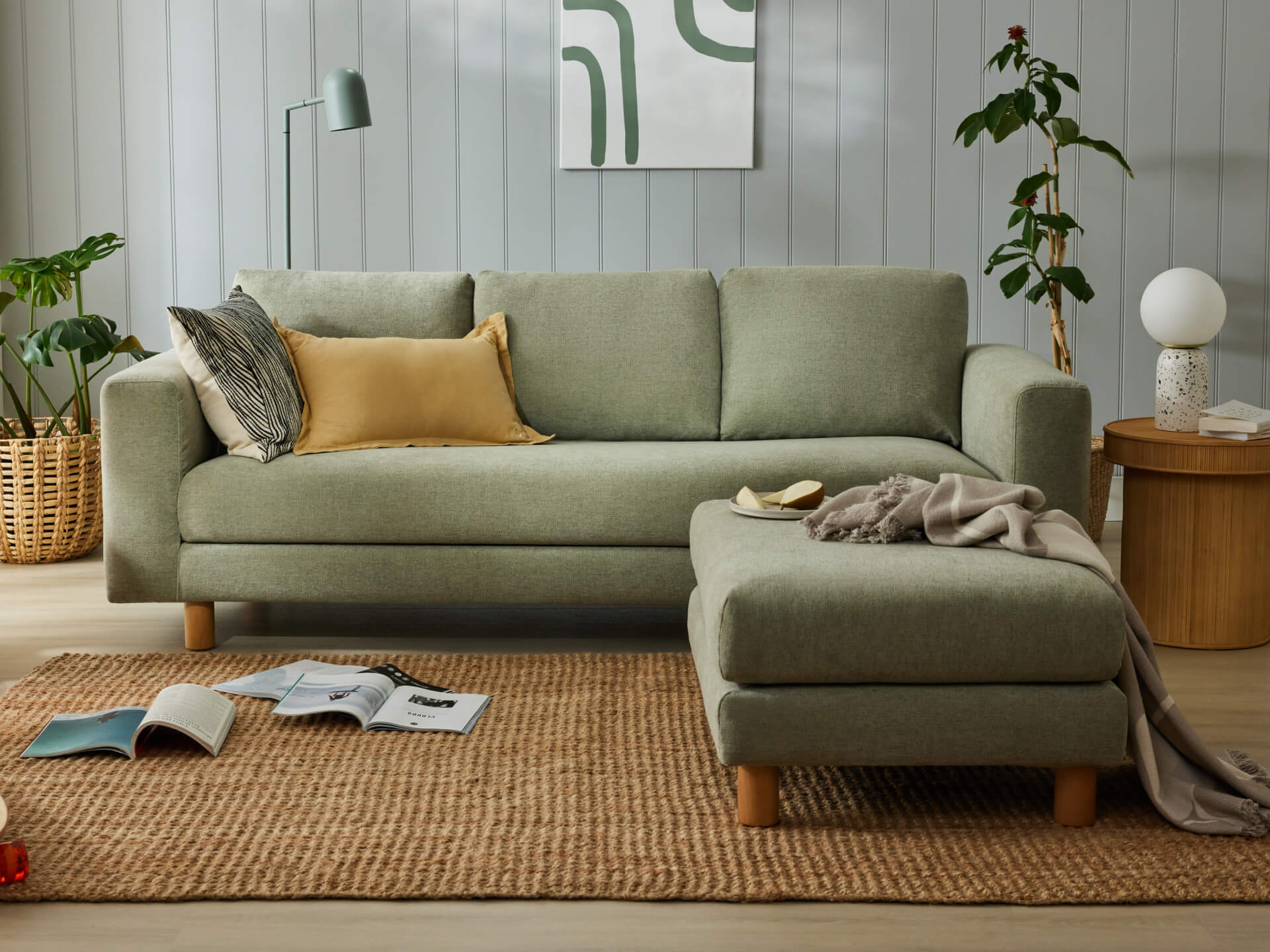 Green three-seater sofa with matching ottoman, topped by cushions and a throw. Set on a natural fibre rug with side table, plants, and wall art.