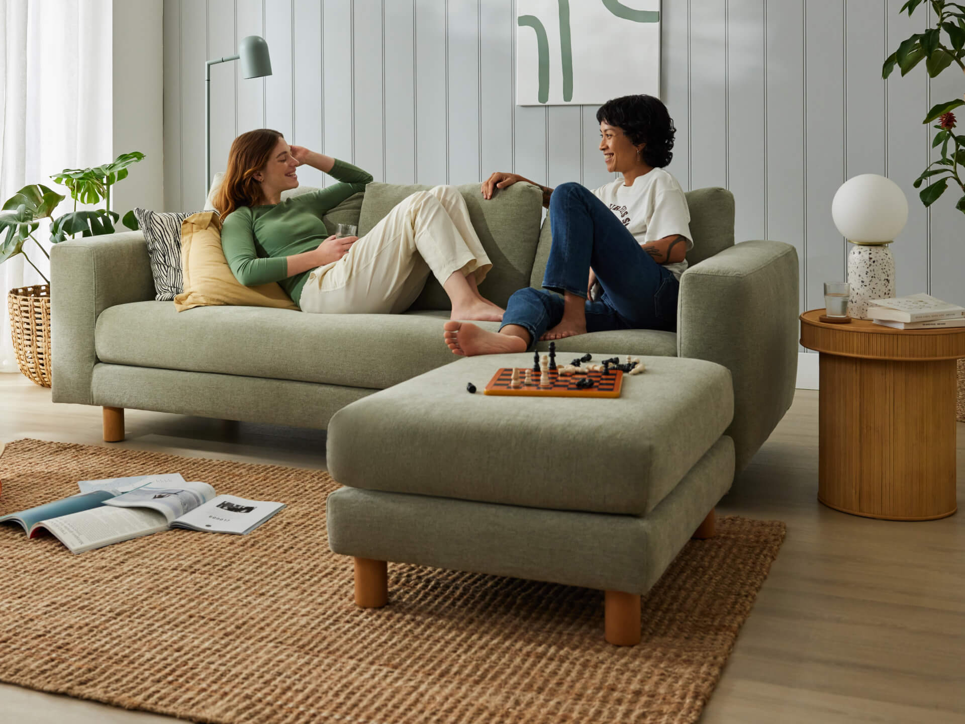 Two people relaxing on a light grey sofa with a matching ottoman. A chess set is on the ottoman. The room has a woven rug and potted plants.