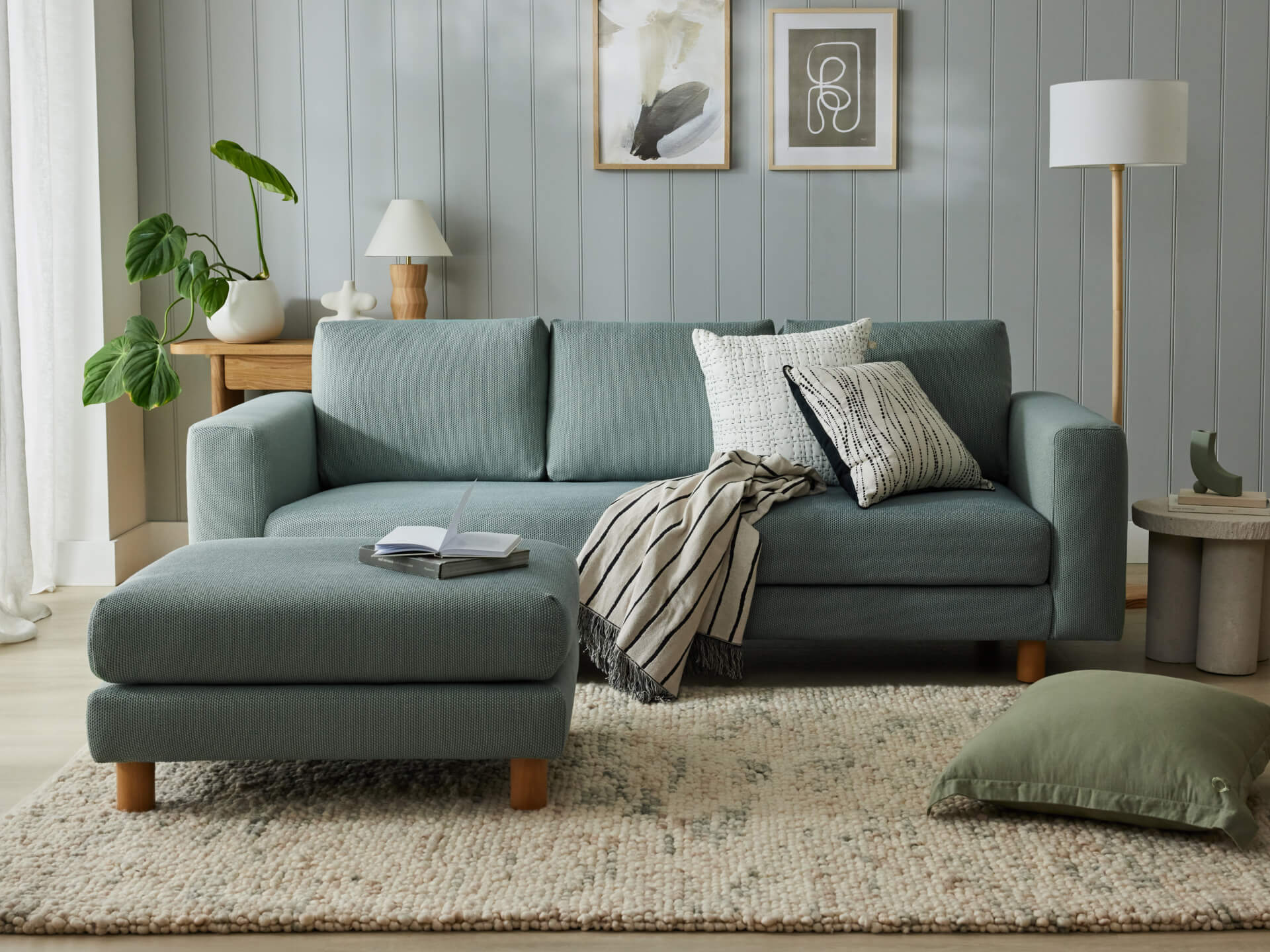Light green sofa with matching ottoman, striped and textured cushions, on beige rug; wood accents, floor lamp, and two abstract art pieces on the wall.