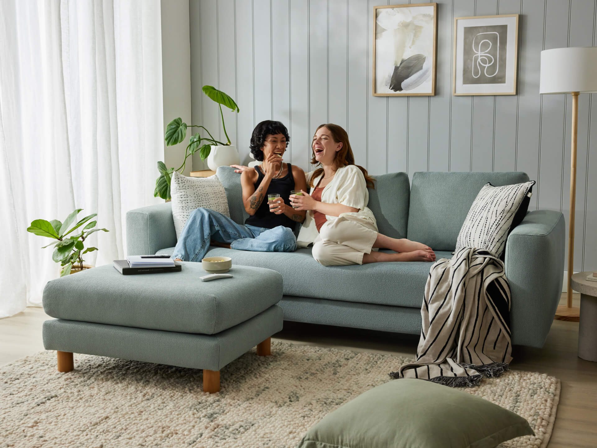 Cosy living room with a light grey sofa, matching ottoman, white and green cushions, potted plants, and framed wall art.