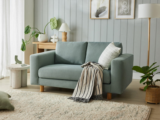 Stylish living room with a light blue sofa, white pillow, and striped throw. Wooden side table with lamp, wall art, and potted plants enhance decor.