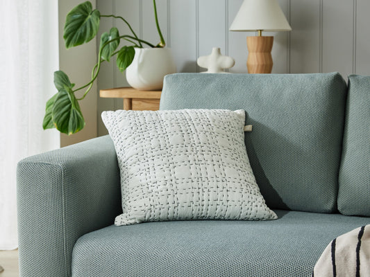Light grey couch with textured cushion, wooden side table, and potted plants. Modern decor with soft lighting and neutral tones.
