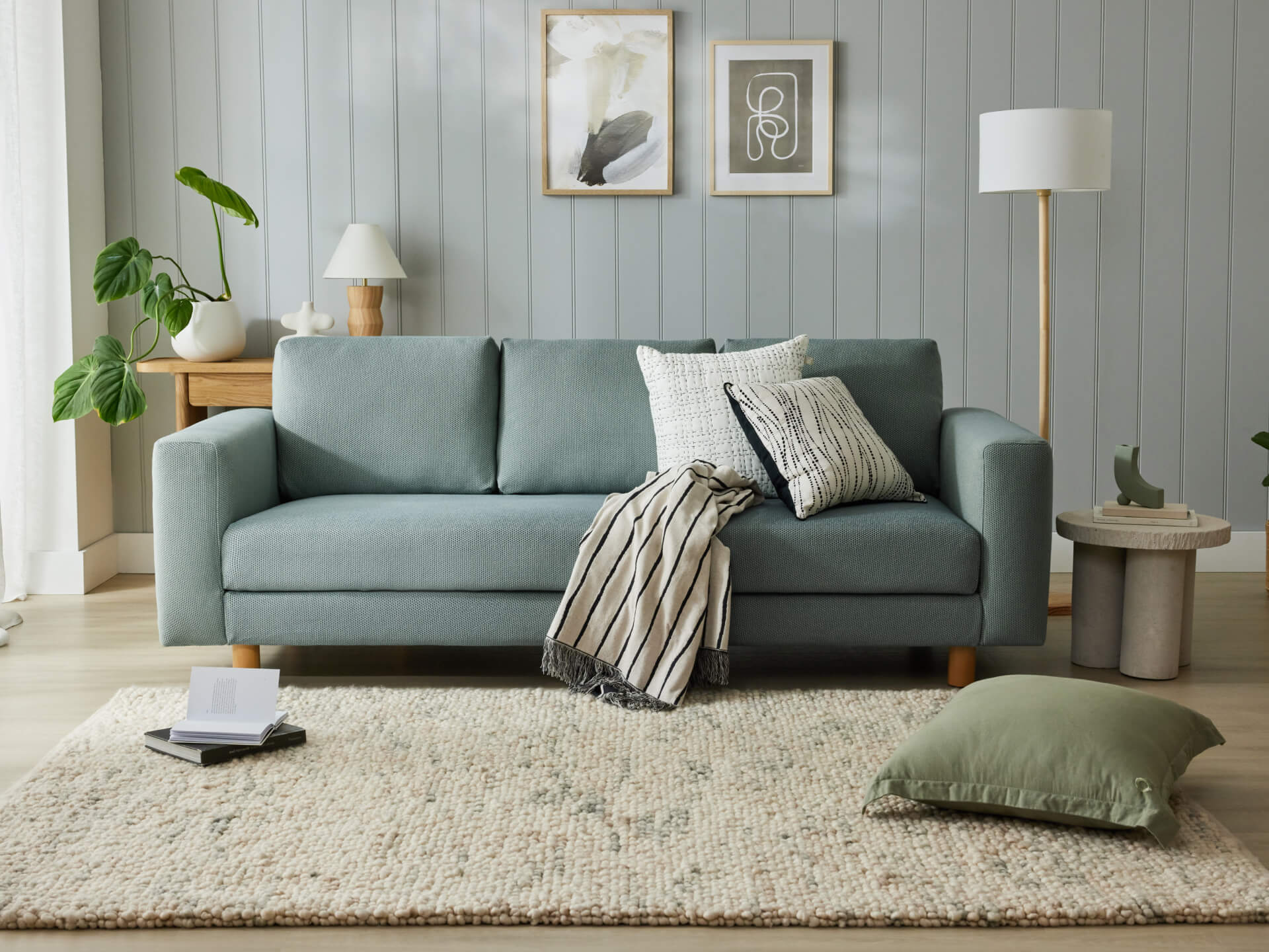 A light blue sofa with cushions and a striped throw, set in a cosy living room with light grey walls, decor, and leafy plants on a soft rug.