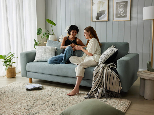 Two people sit on a light grey couch in a cosy living room with cushions and plants. A soft rug and decorative wall art complete the setting.