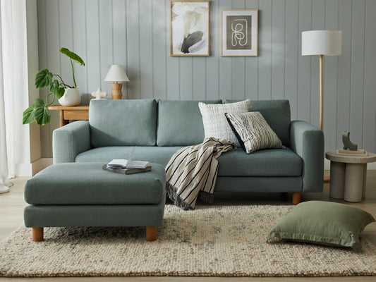 Light grey couch with wooden legs, adorned with cushions and throw, next to matching ottoman on a beige rug. Framed art and potted plant nearby.