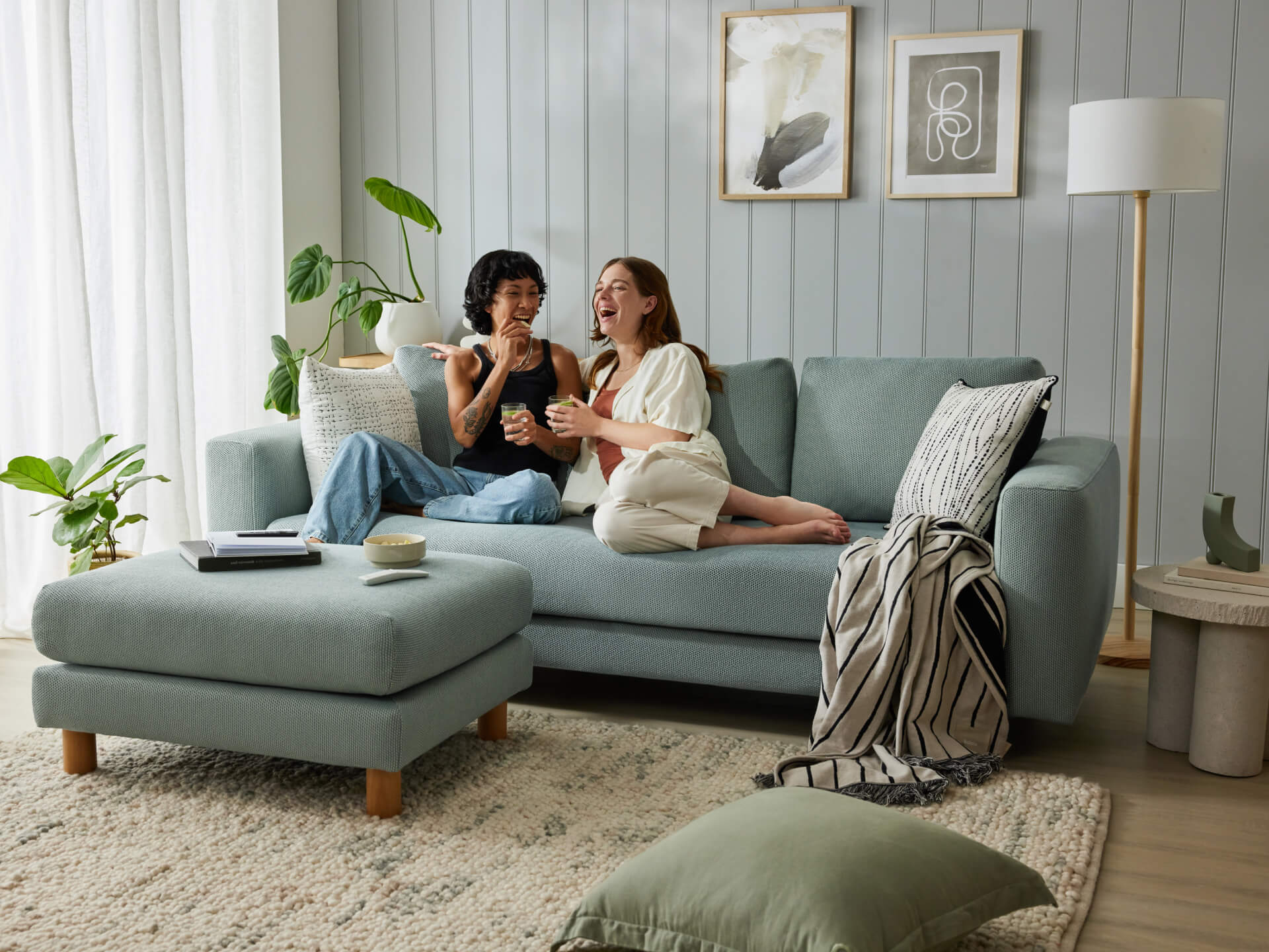 A light grey sofa with a matching ottoman, wooden legs, and assorted cushions. Nearby, there's a large potted plant, framed art, and a floor lamp.
