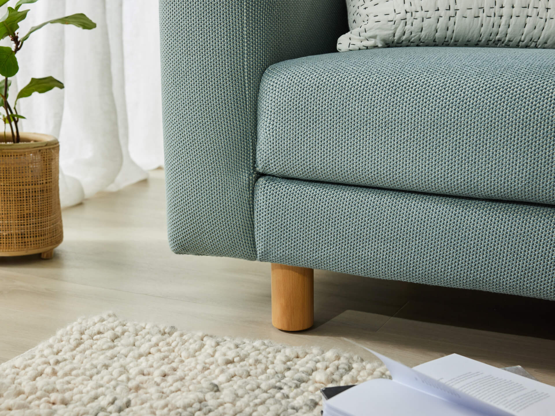 Close-up of a light grey upholstered couch with wooden legs on a light wood floor. A cream rug, pot plant, and a book are nearby.