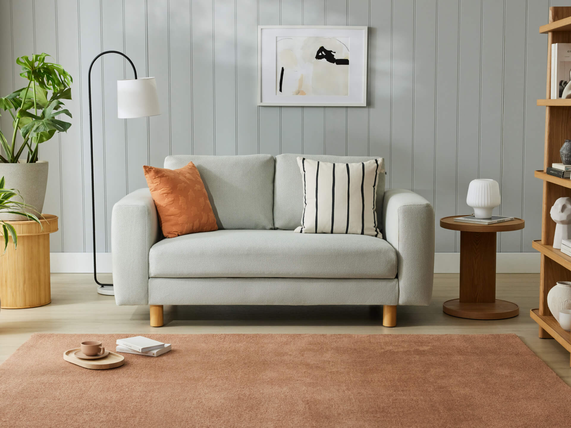 Light grey sofa with orange and striped cushions, tan rug, wooden side table, floor lamp, plants, and abstract wall art in a modern living room.