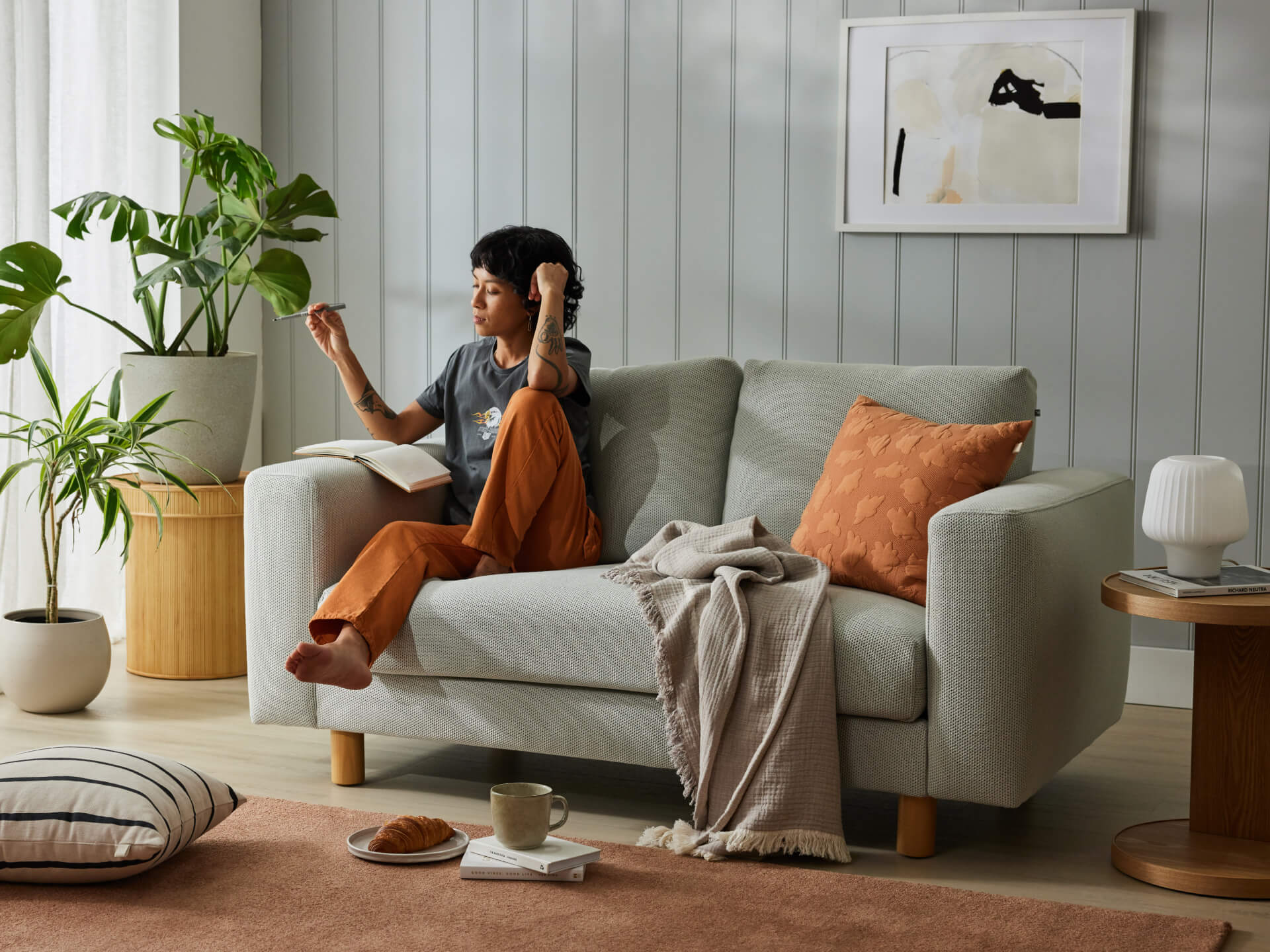 A person sits on a light grey sofa with orange cushions and a beige throw. Nearby are houseplants, a wooden side table, and minimalistic décor.