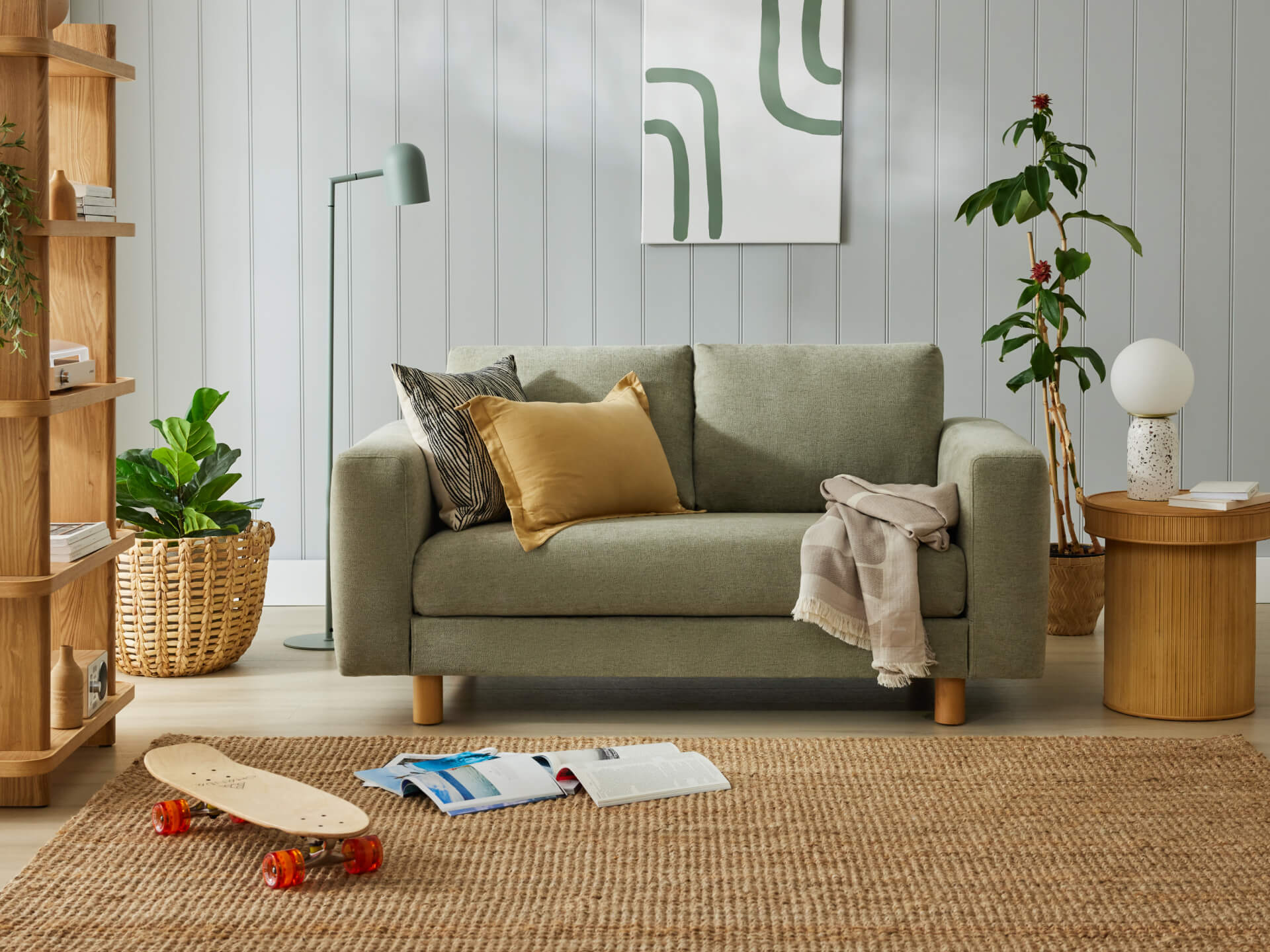 Sage green sofa with mustard and black cushions, woven rug, lamp, skateboard, plants, and art on light grey panelled wall. Cosy living room.
