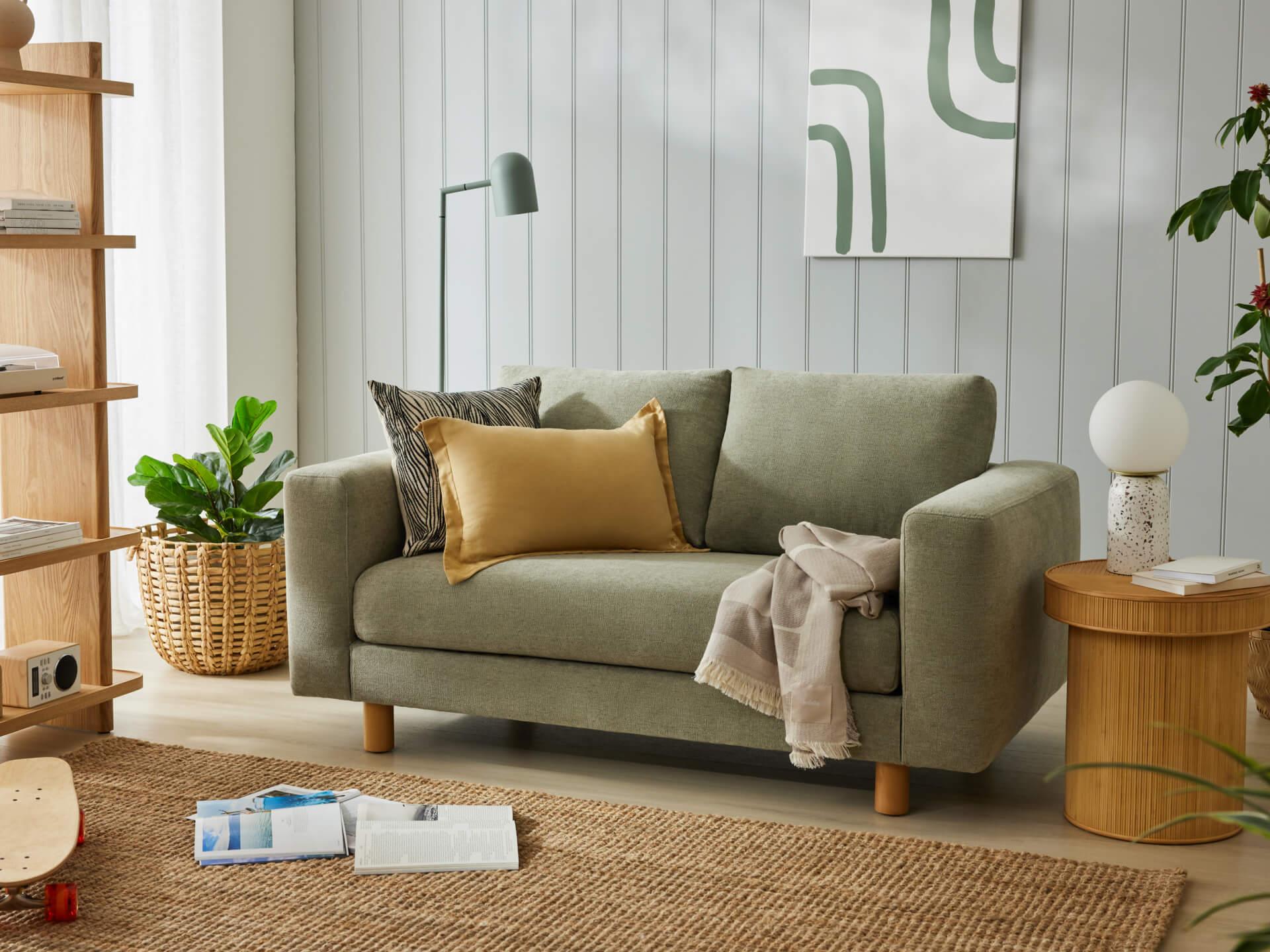A small light grey sofa with mustard and patterned cushions, a throw blanket, surrounded by indoor plants and a wooden side table.