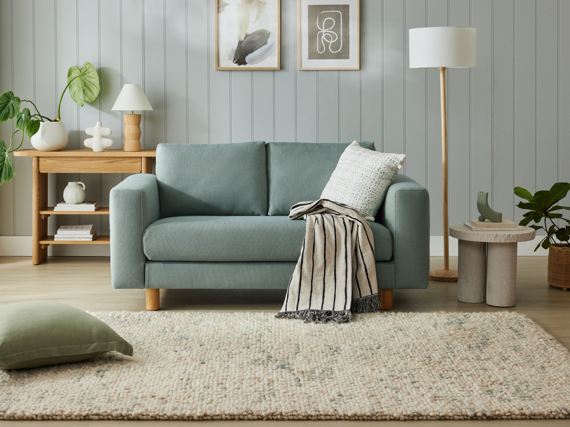 Cosy living room with a light blue sofa, striped throw, white cushion, wooden lamp, side table, and plants. Rug on light wood floor.