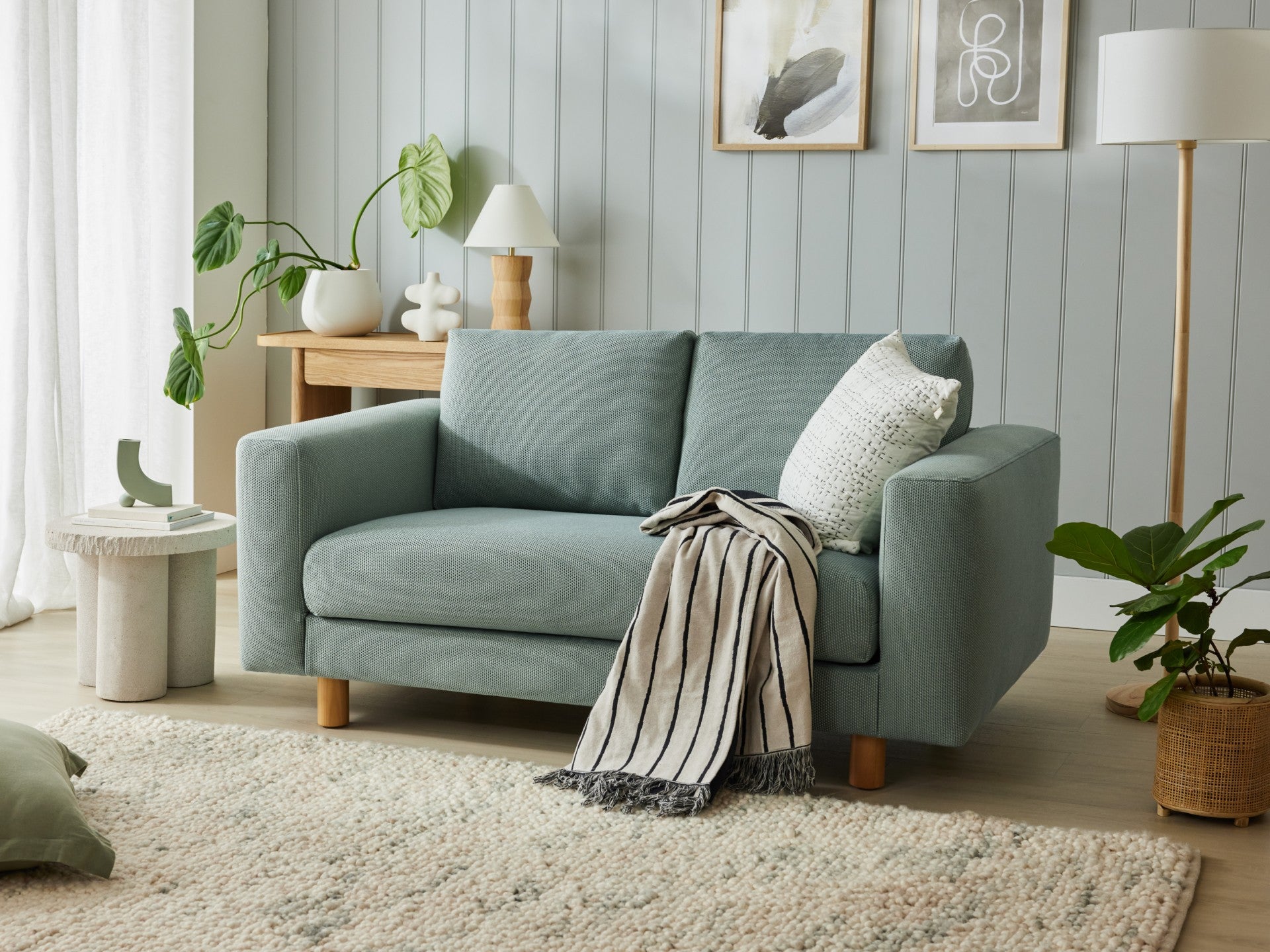 Light green sofa with wooden legs, striped throw, and cushion. Styled with indoor plants, side tables, and wall art in a cosy living room.