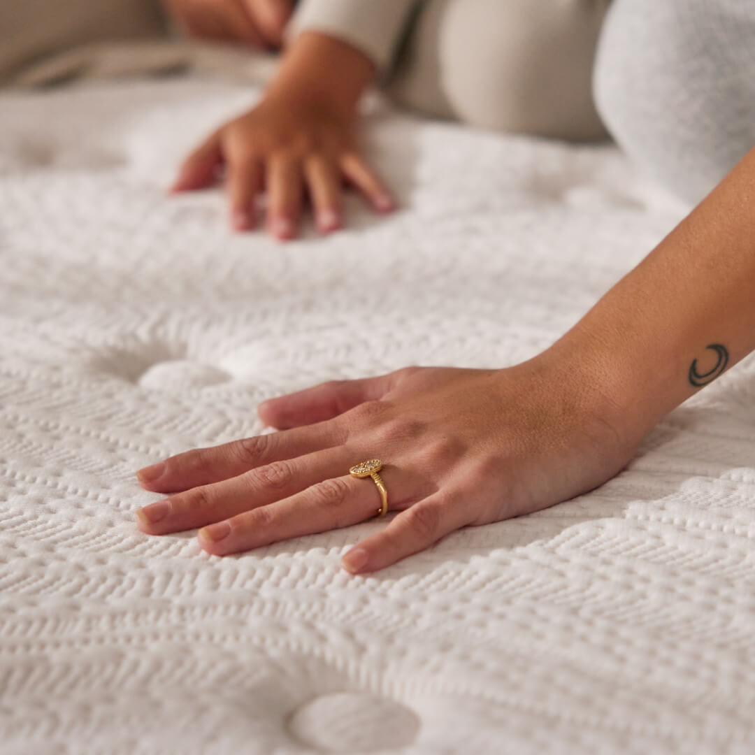 Hand with a gold ring pressing on a textured white mattress surface, showcasing its softness. Another hand is visible in the background.