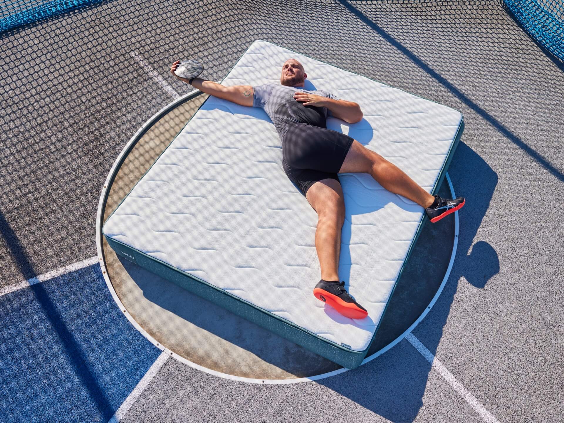 Man in athletic wear relaxes on a light grey mattress set on an outdoor trampoline. Bright sunlight casts shadows on the textured surface.