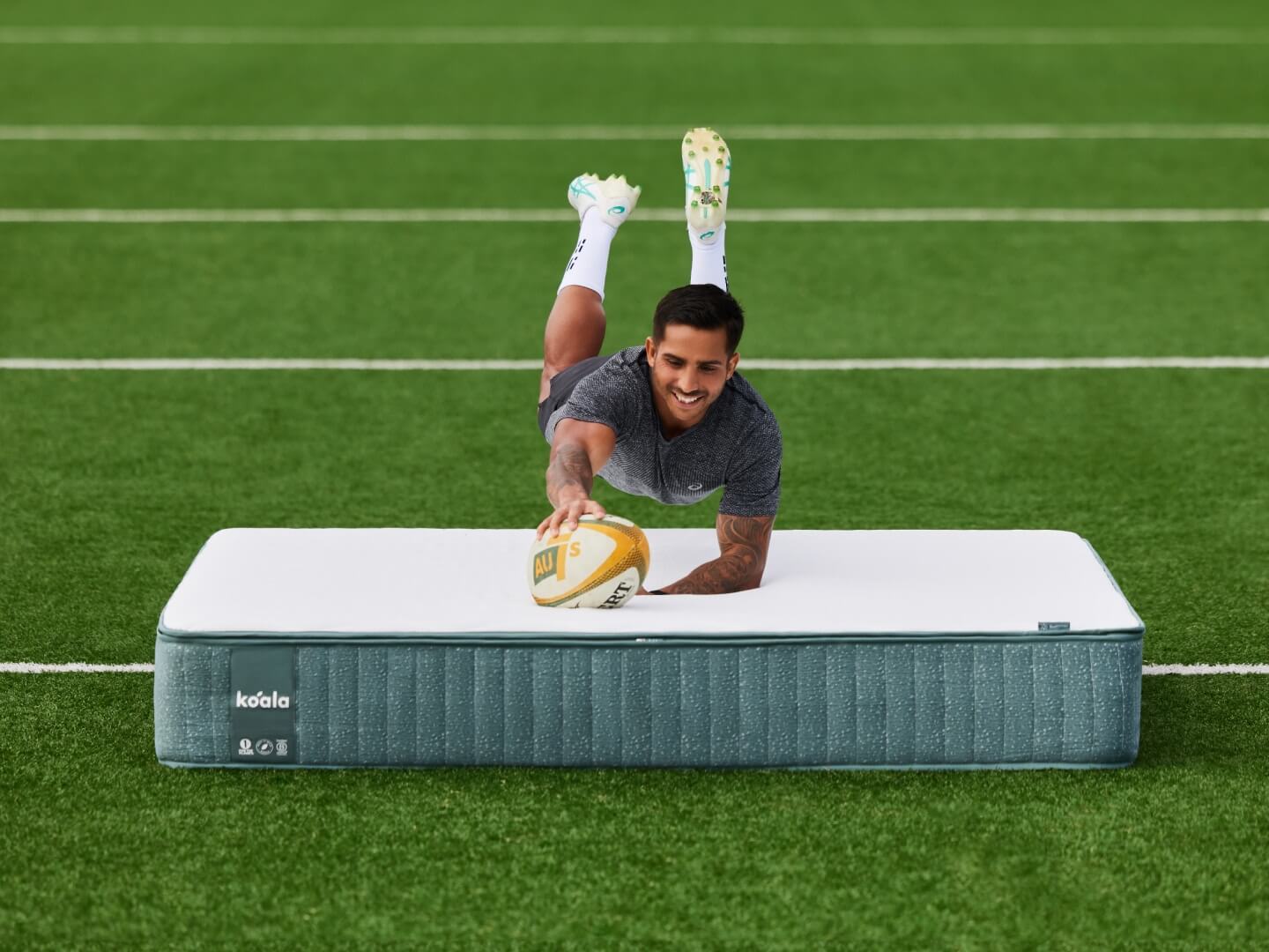 Person diving onto a white and grey mattress on a grass field, holding a rugby ball.