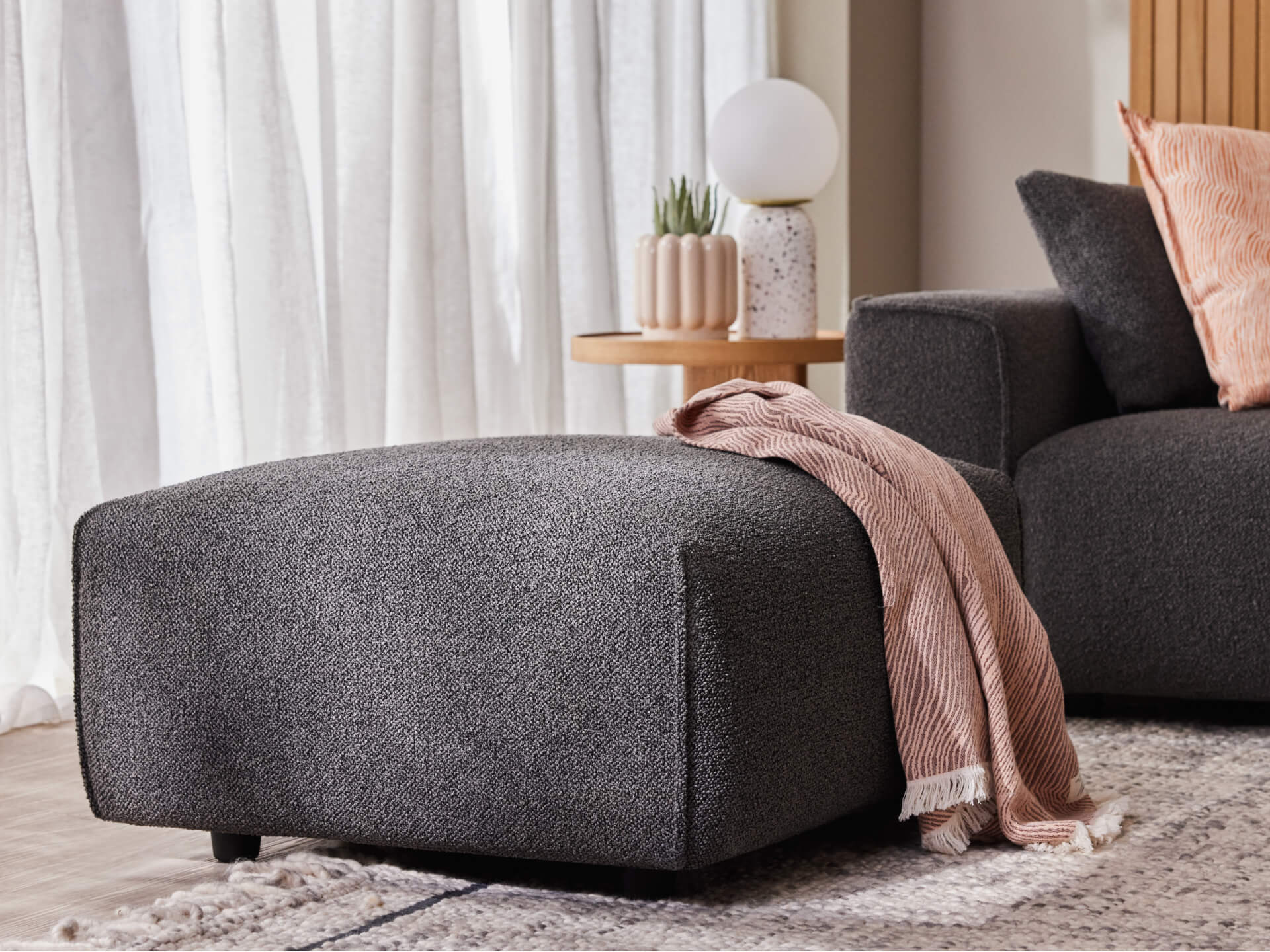 Dark grey ottoman with pink throw beside a matching sofa. A wooden side table with a lamp and plants is in the background near white curtains.