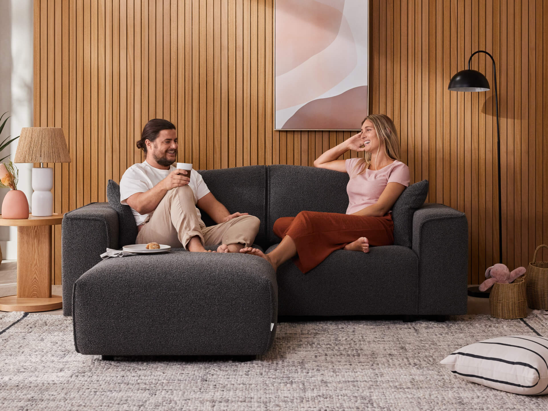 Couple relaxing on dark grey sofa with matching ottoman in cosy living room. Wooden panel wall, modern lamp, and plush rug create a warm atmosphere.